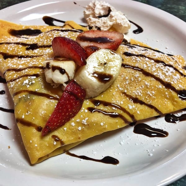 Golden crepe drizzled with chocolate, powdered sugar, strawberries, bananas, and whipped cream on a white plate.