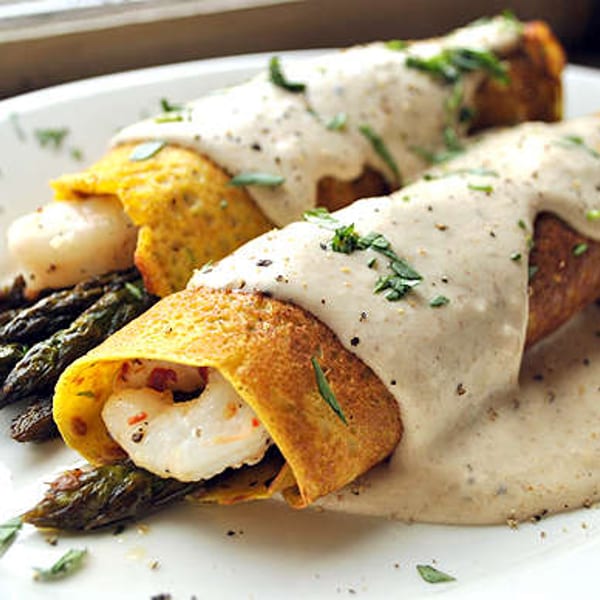 Two shrimp and asparagus crepes covered in creamy sauce and fresh herbs on a white plate.