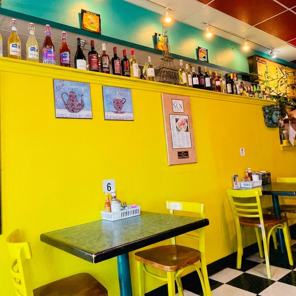 Vibrant yellow restaurant interior with checkered floors, tables, chairs, and bottles lining a shelf.