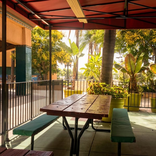 Outdoor patio with a wooden picnic table and benches under a red awning, surrounded by lush potted plants.