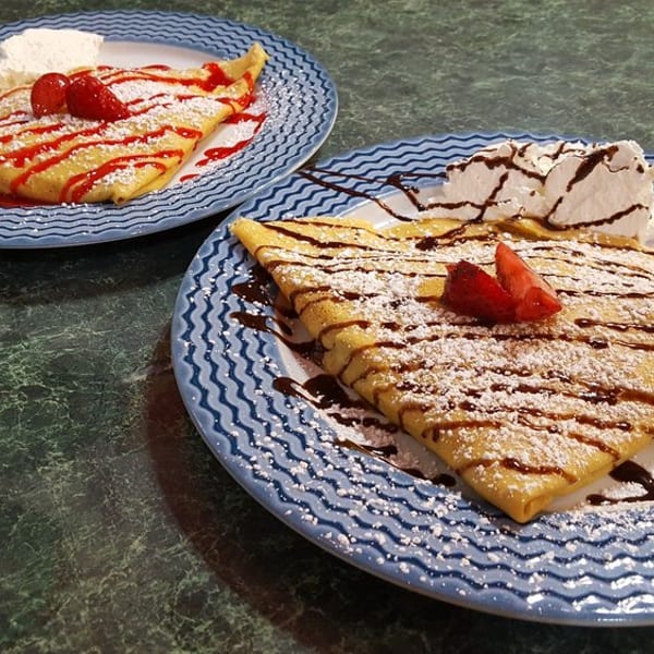 Two dessert crepes on blue plates, one with strawberry sauce, the other with chocolate, both with whipped cream.