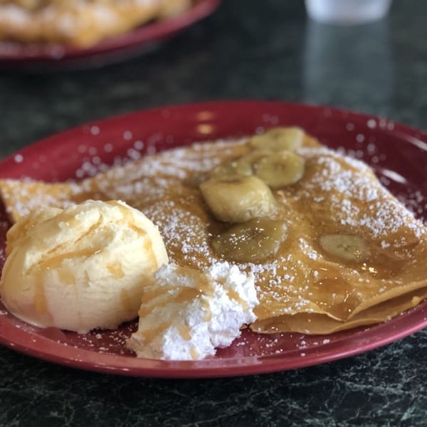 Banana crepe with powdered sugar, whipped cream, and a scoop of vanilla ice cream on a red plate.