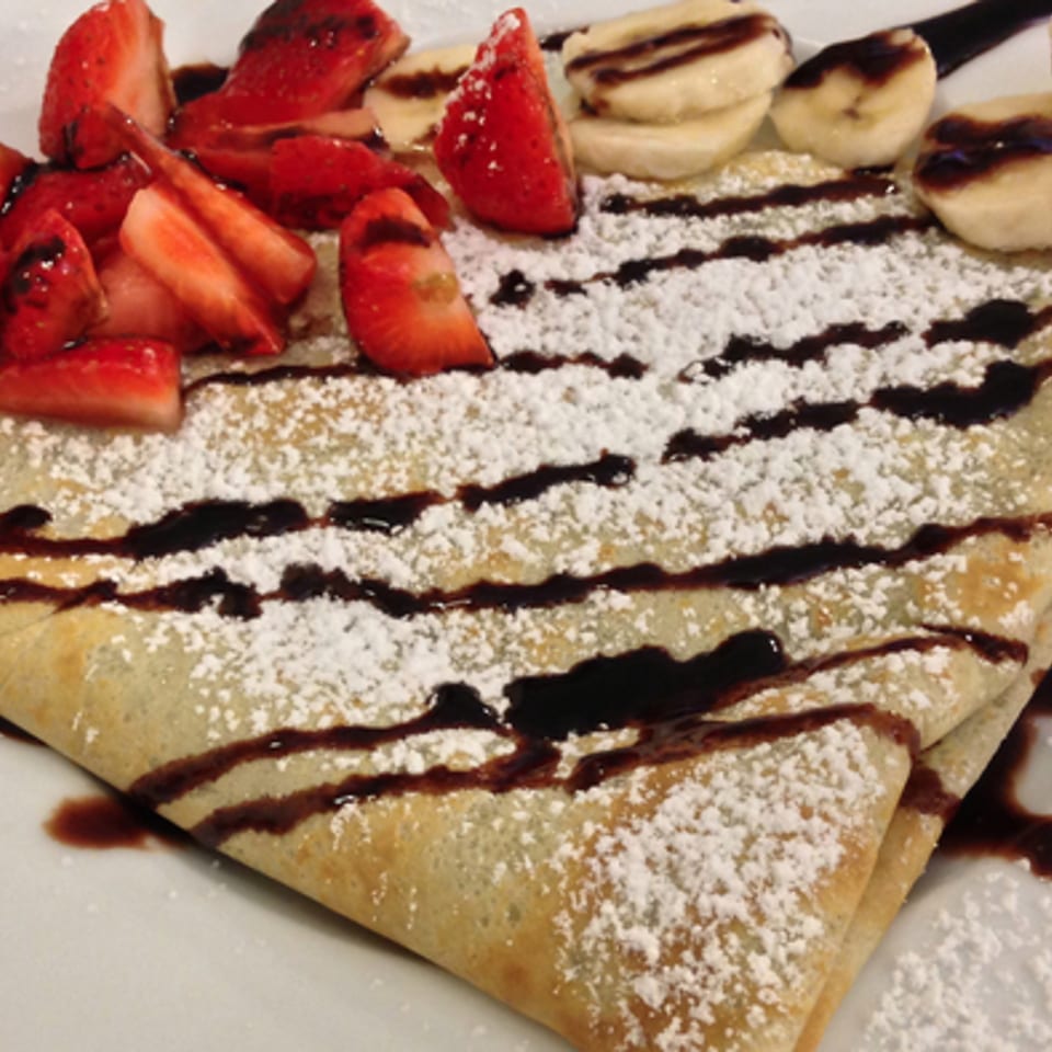 A folded crepe topped with sliced strawberries, bananas, chocolate drizzle, and powdered sugar.