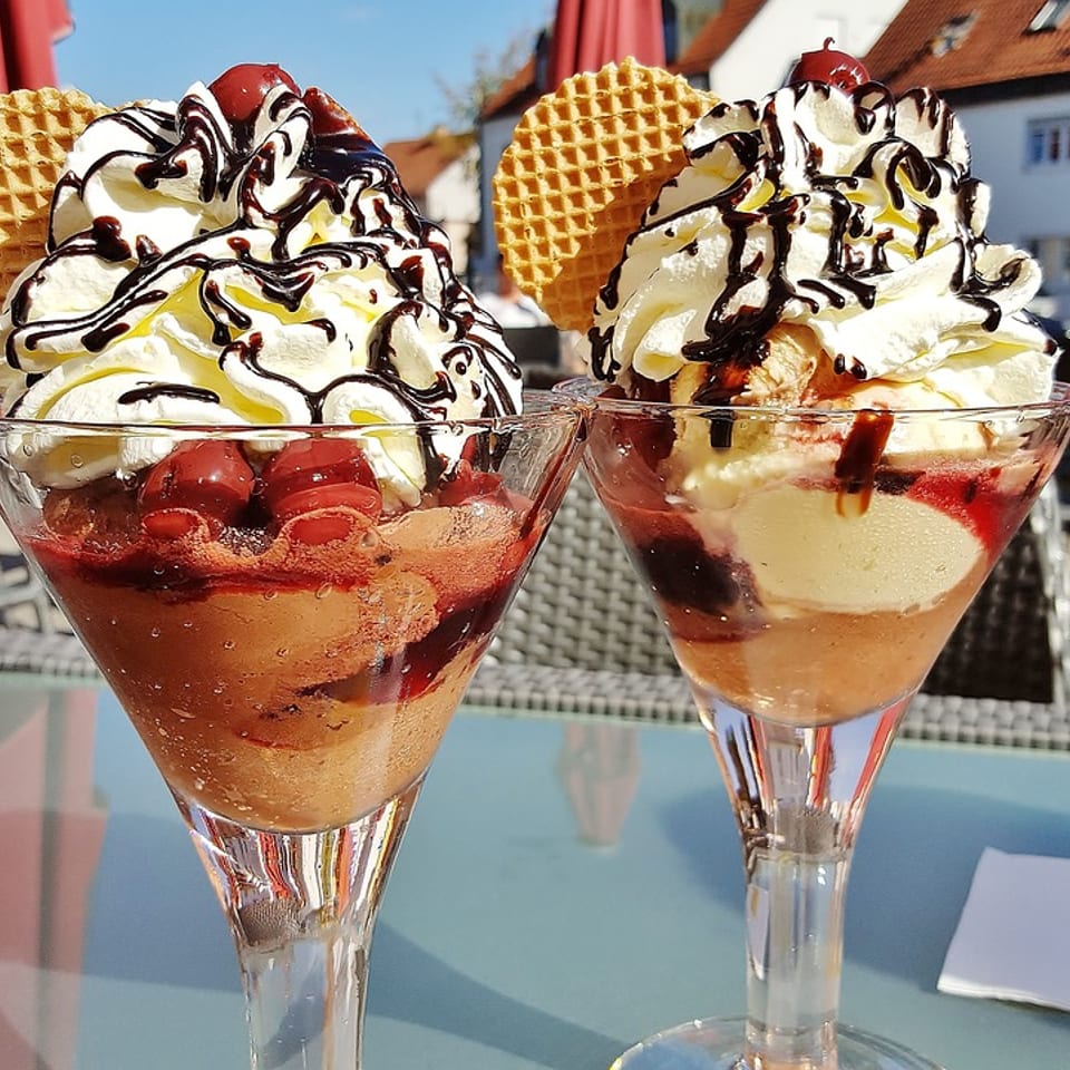 Two ice cream sundaes with whipped cream, chocolate sauce, cherries, and waffle cookies on an outdoor table.